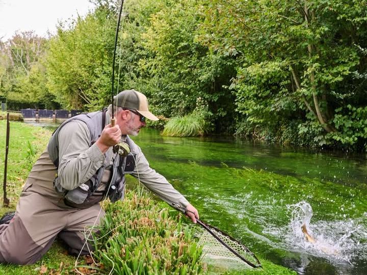 Ben Bangham kneeling on a chalk stream bank, playing a trout